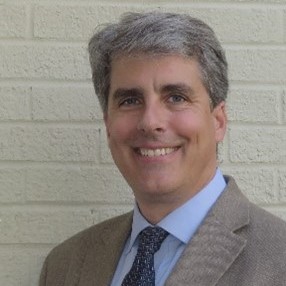Photo of Doug Bellomo. Photo shows a smiling man from the shoulders up standing in front of a white brick wall. The man has short grey hair, a tan suit, light blue shirt, and a dark blue patterned tie.