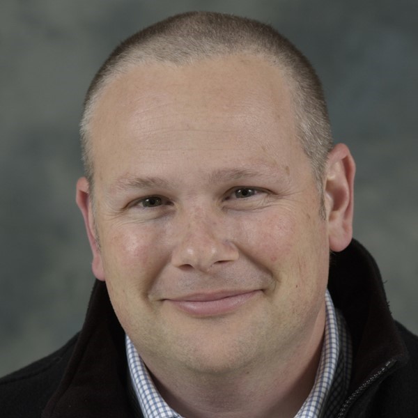 Photo of Dr Rob Young. Photo shows a headshot of a smiling man with short cropped grey hair, a blue gingham shirt, and a black jacket in front of a grey background. 